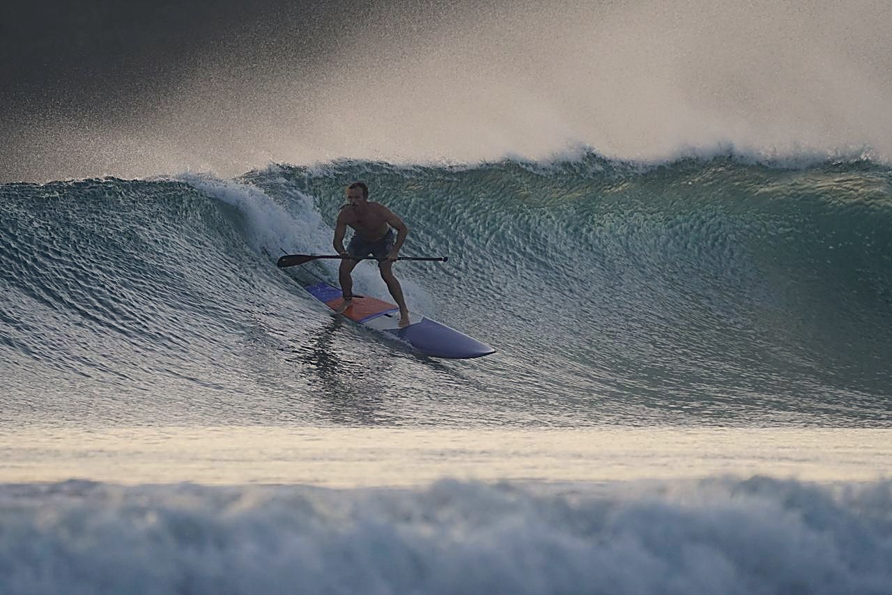 Dr. Aaron Burt dropping into a wave on a stand-up paddle board, backlit at dusk